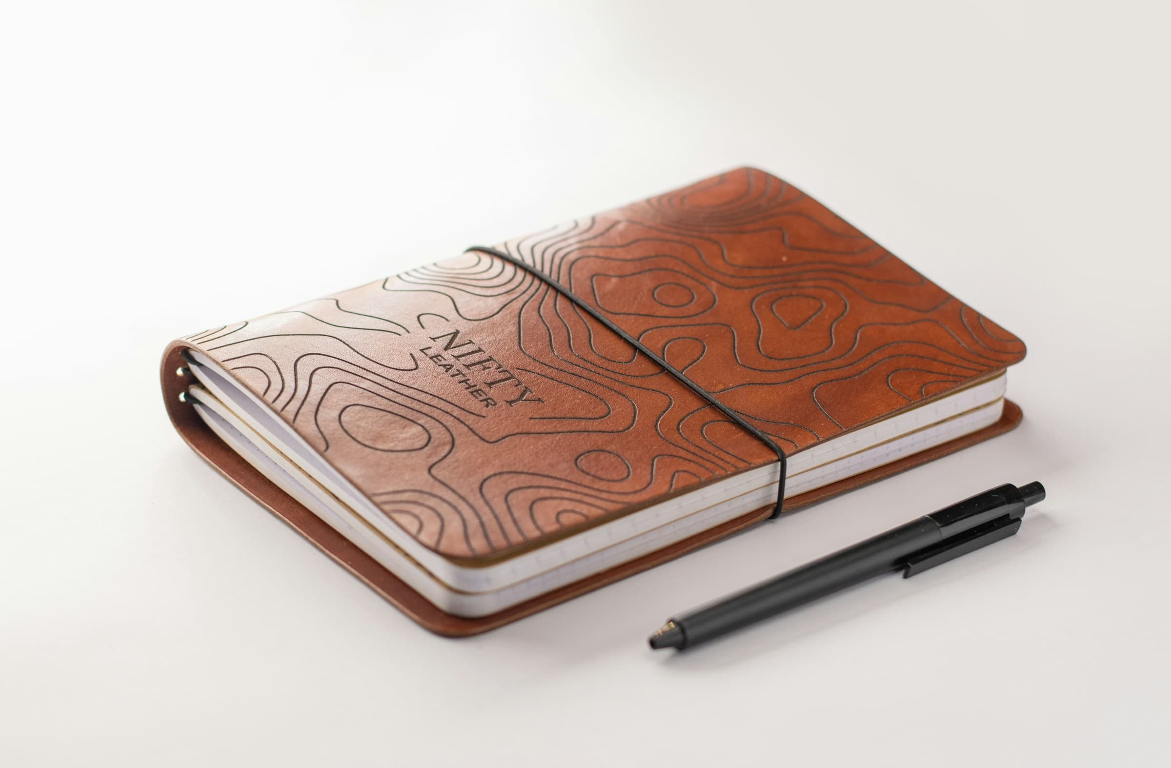 A leather field journal with topographic embossing, alongside a black pen on a clean surface — editorial reference for the documentary nature of the journal.